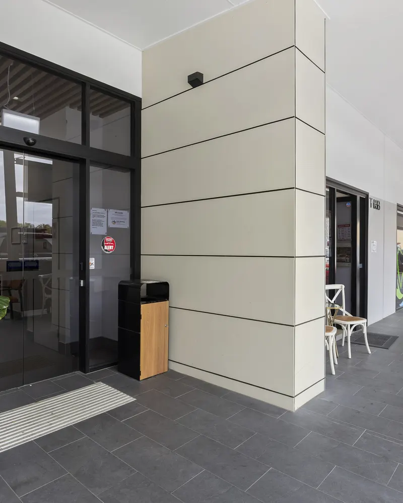 Automatic glass entrance doors off a covered, tiled walkway, with café chairs along the walkway
