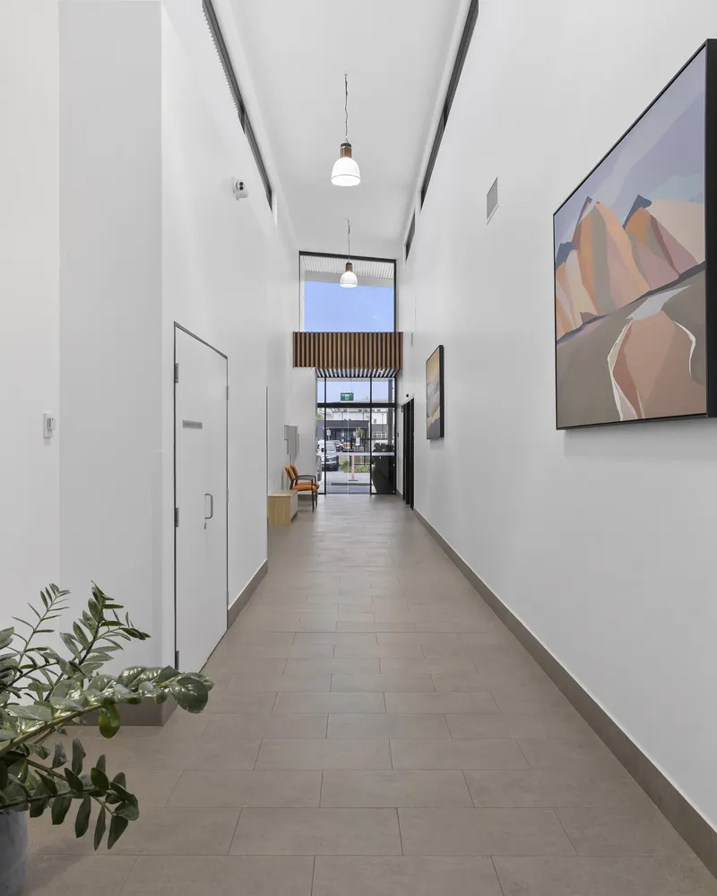 A tall, tiled corridor with pendant lights, high windows, artwork on the walls and indoor plants, leading to the glass entrance