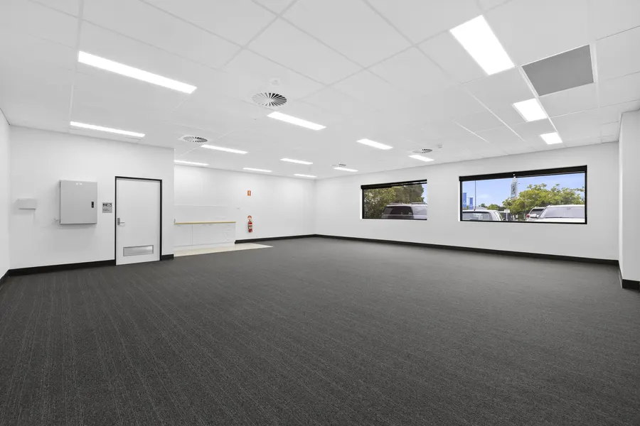 An empty open-plan suite with charcoal carpet, a suspended ceiling with LED panels and air-conditioning outlets, a kitchenette bench and two windows looking out to the car park